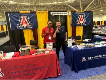 Two men giving the Wildcat hand gesture standing behind conference booths for the Haury Program and Community Engagement and Partnerships.